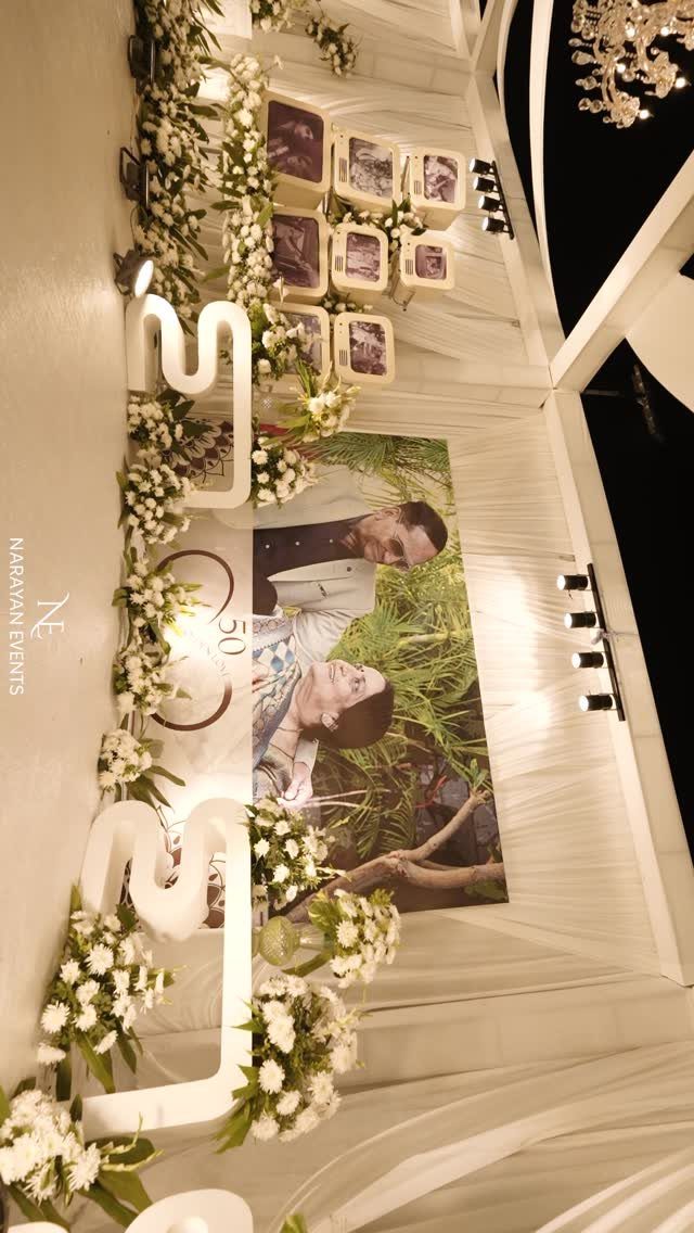 Celebratory 50th anniversary backdrop with white flowers, framed photos, and large illuminated numbers.