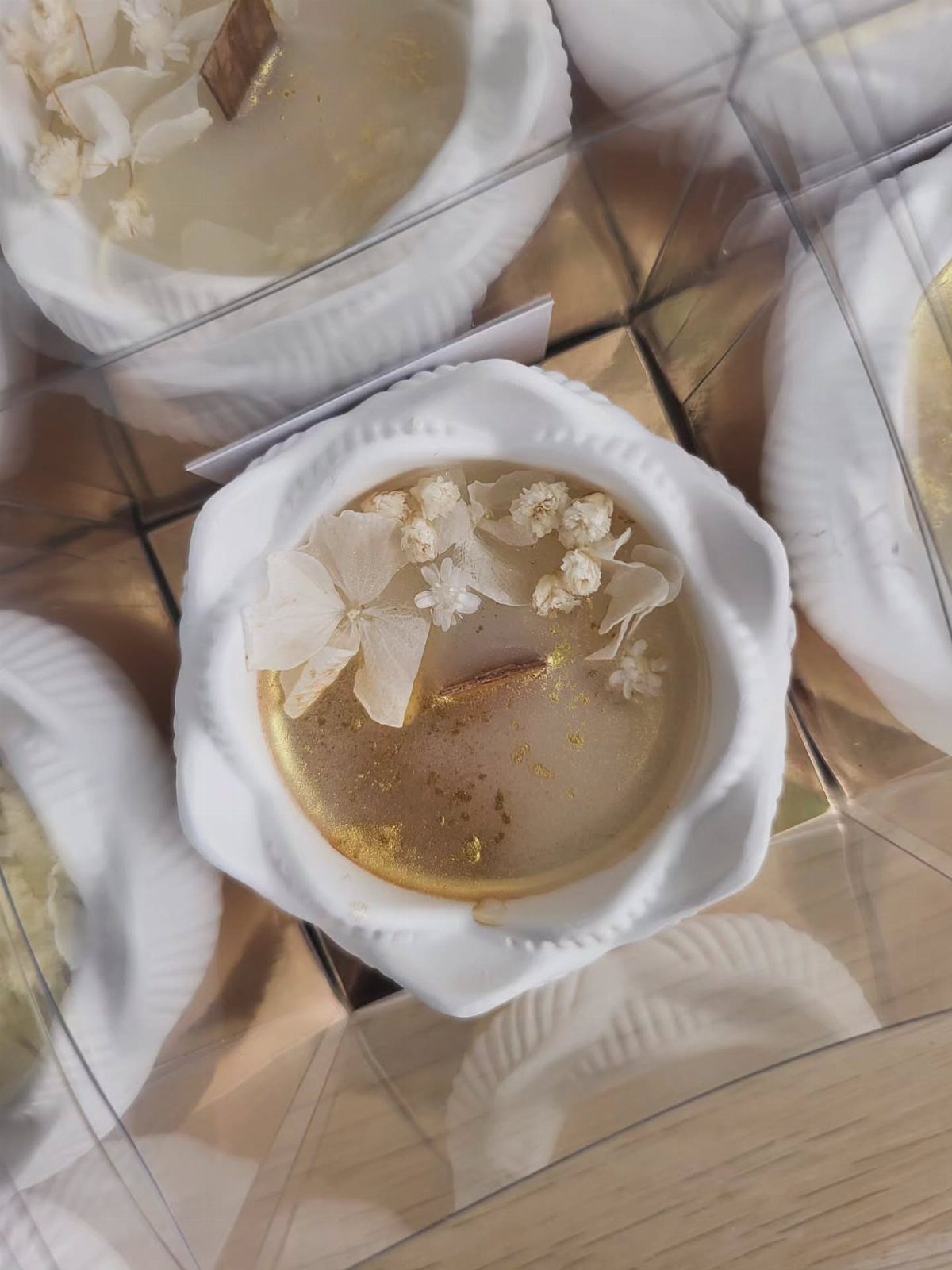 Gold-colored wax candle in a white flower-shaped holder topped with dried white flowers.
