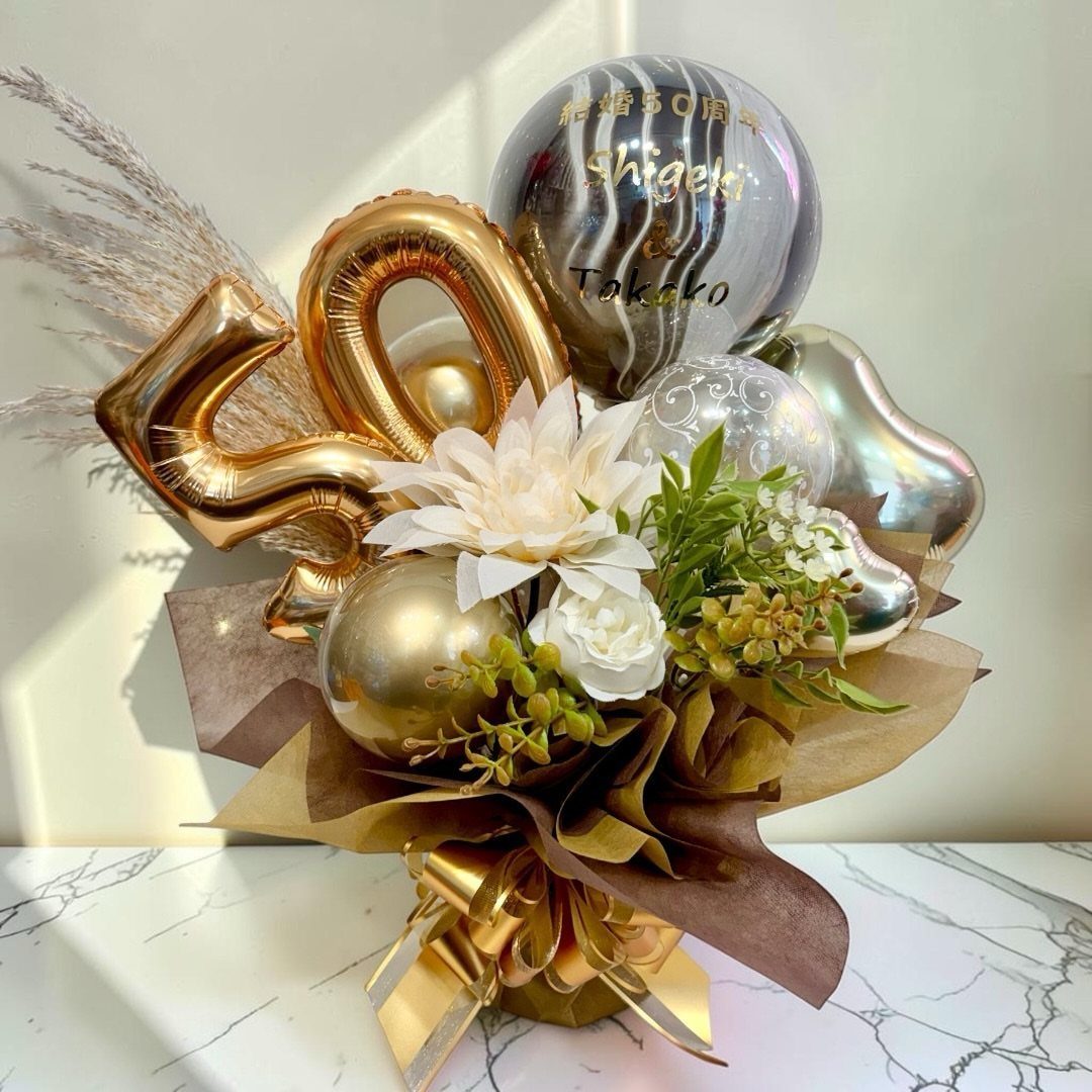 Celebratory bouquet with gold 50 balloons, white flowers, and gold ornaments on marble.