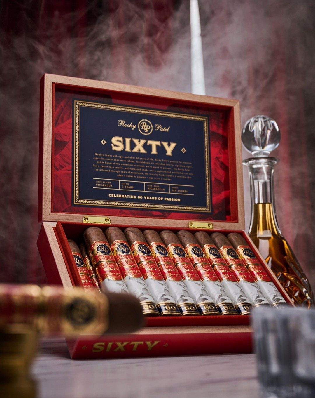 Open wooden cigar box filled with cigars and a 'SIXTY' label, beside a decanter.