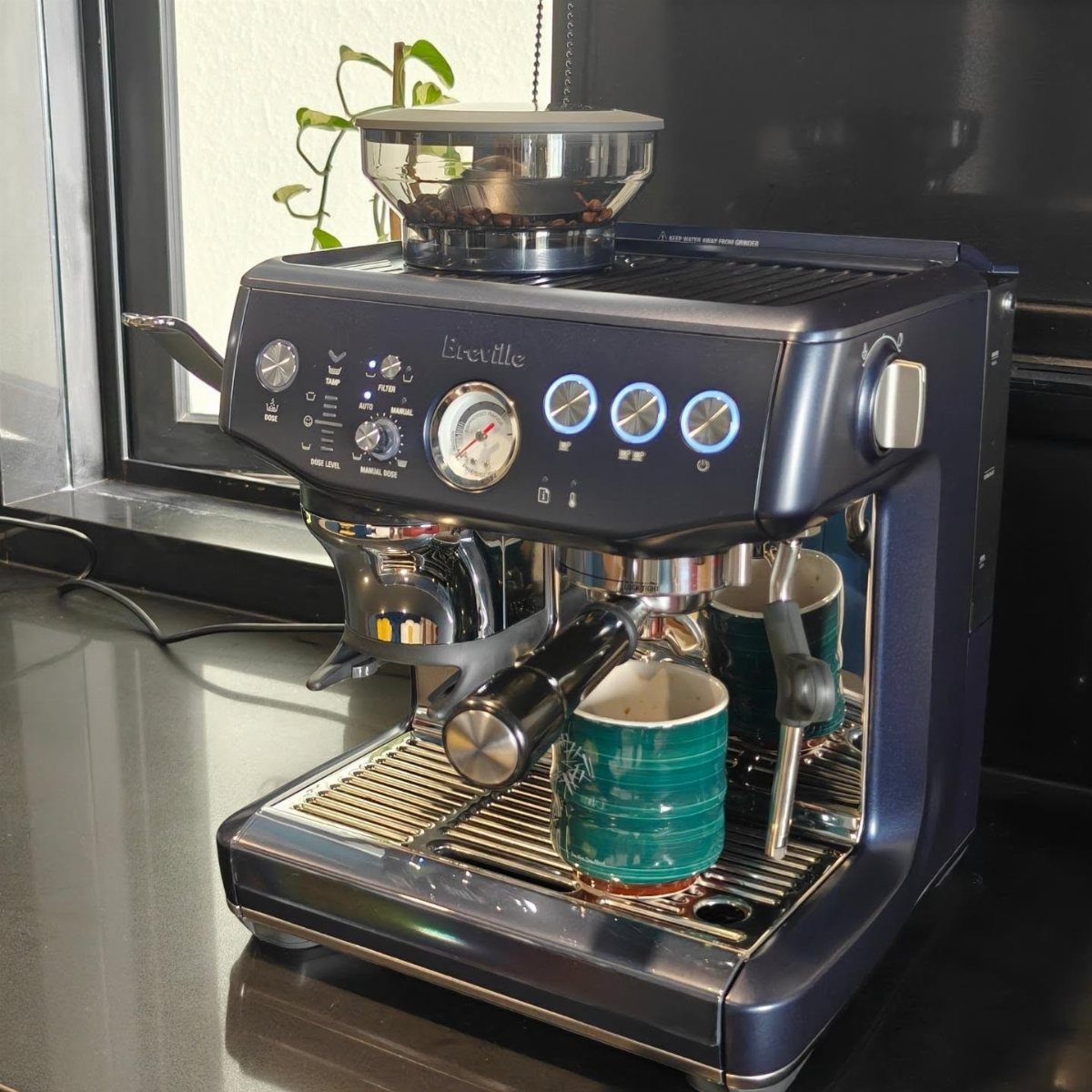 Metallic blue espresso machine with two teal mugs on a dark kitchen counter