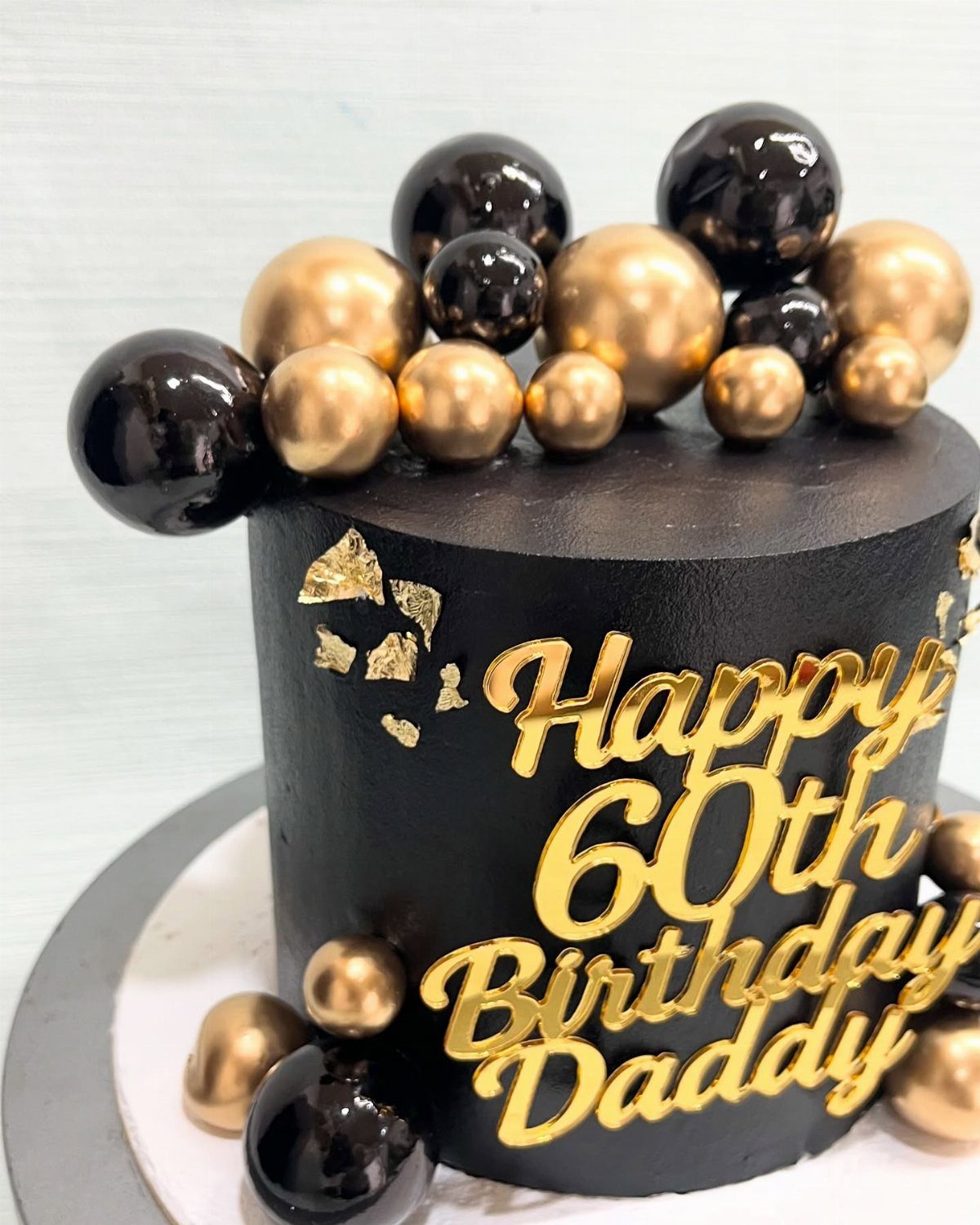 Black birthday cake with gold lettering saying Happy 60th Birthday Daddy and gold spheres.
