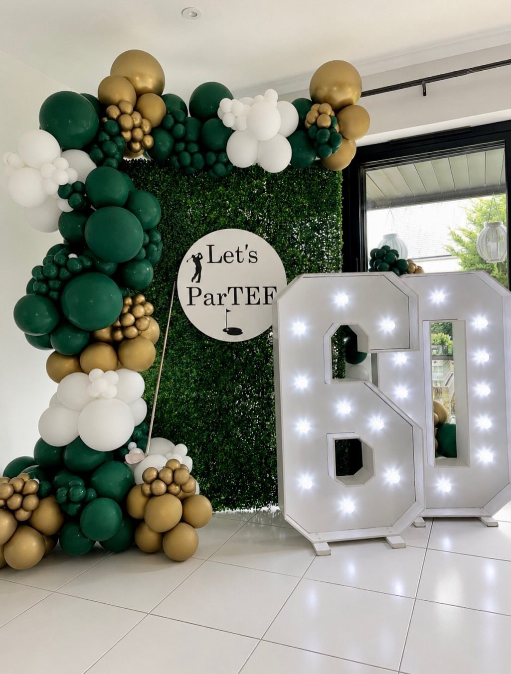 60th birthday party decorations with green balloons, hedge wall, and large lit numbers.