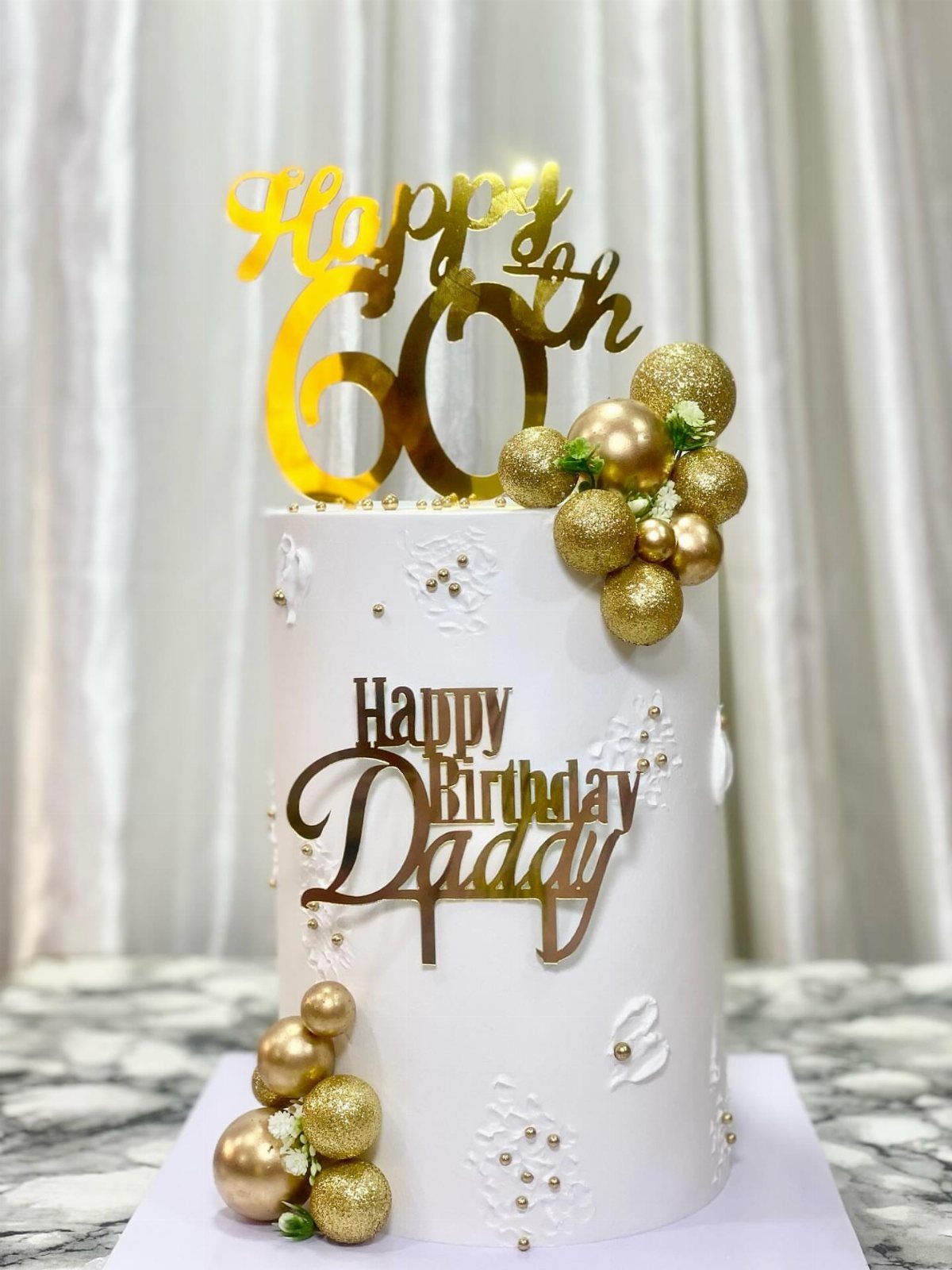 White birthday cake with gold spheres and toppers reading Happy 60th Birthday Daddy