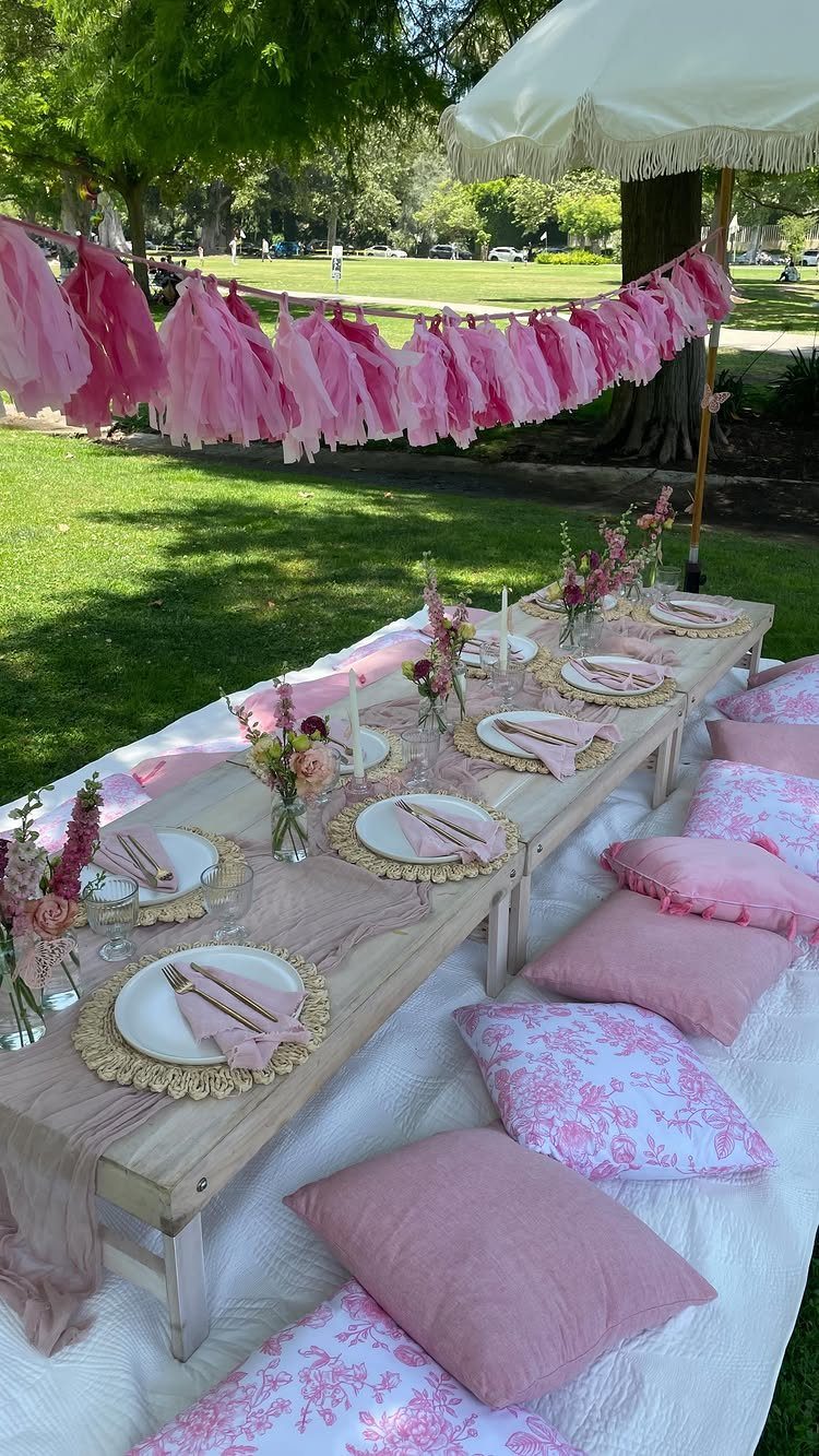 Outdoor picnic party setup with a low wooden table, pink decorations, and pillows.