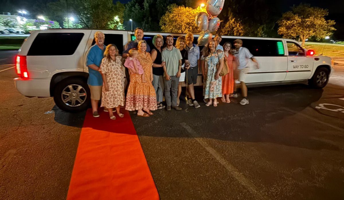 Group of adults posing by a white stretch limousine on a red carpet.