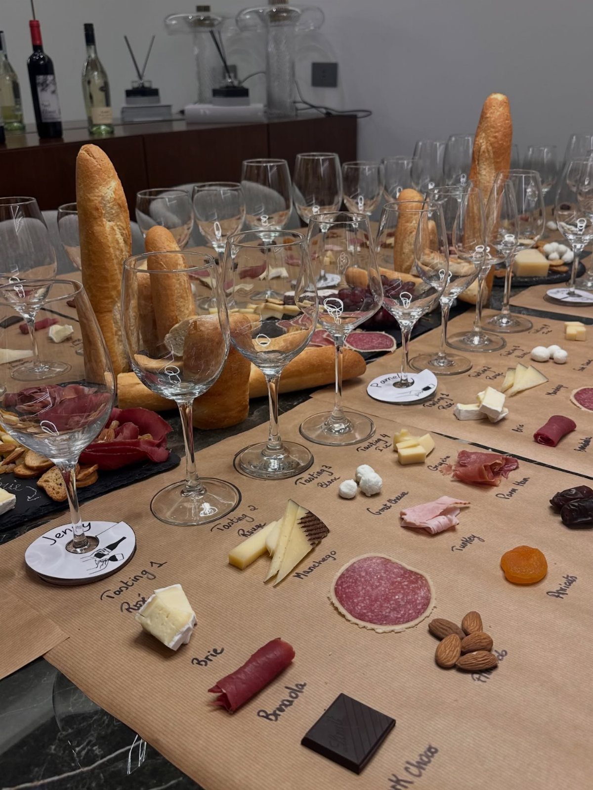 Wine tasting table spread with glasses, baguettes, cheeses, meats, and chocolate on brown paper.