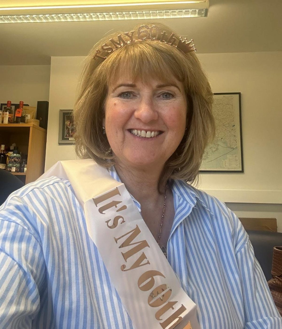 Smiling woman wearing a 60th birthday sash and gold crown indoors.