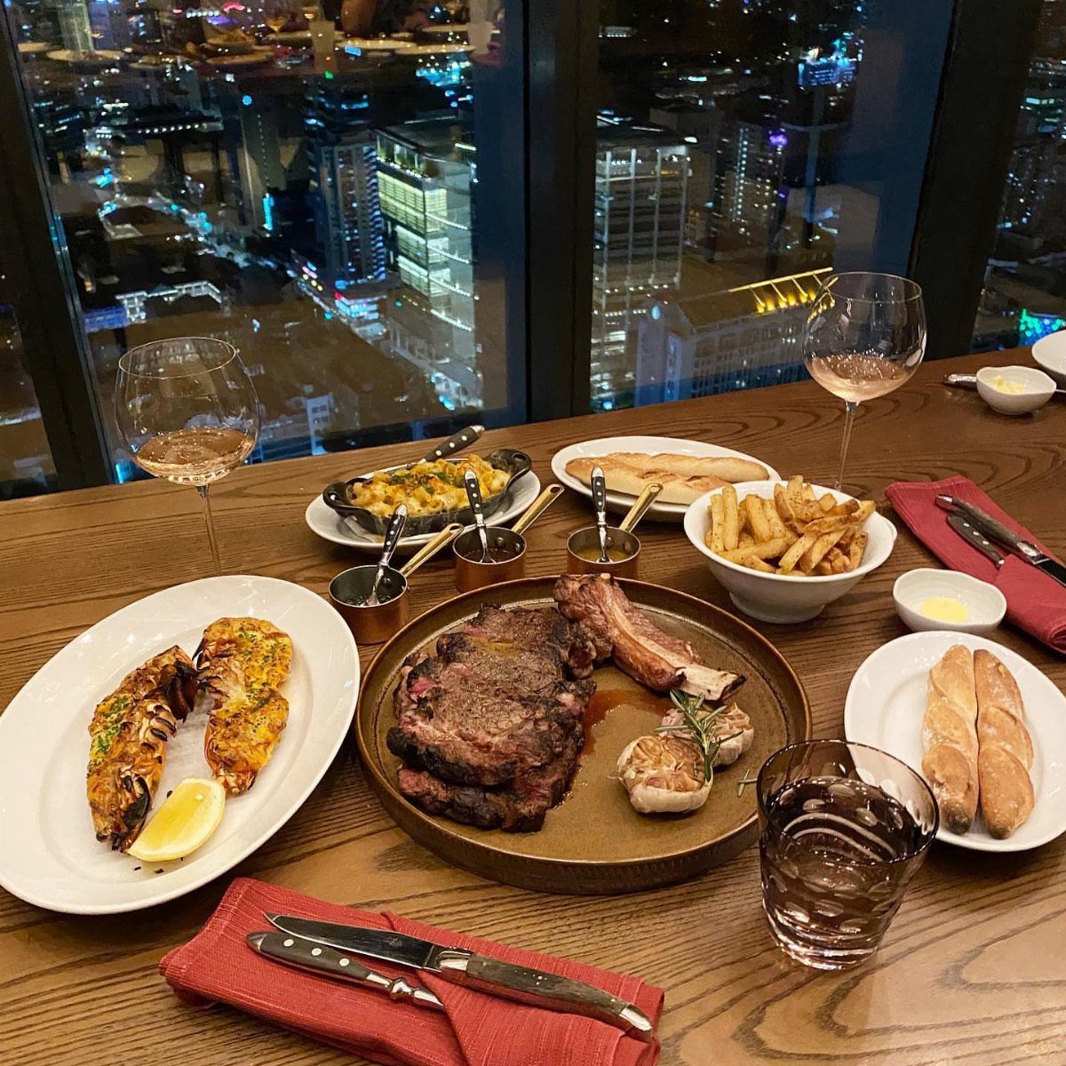 Gourmet dinner spread on a wooden table overlooking a city skyline at night.