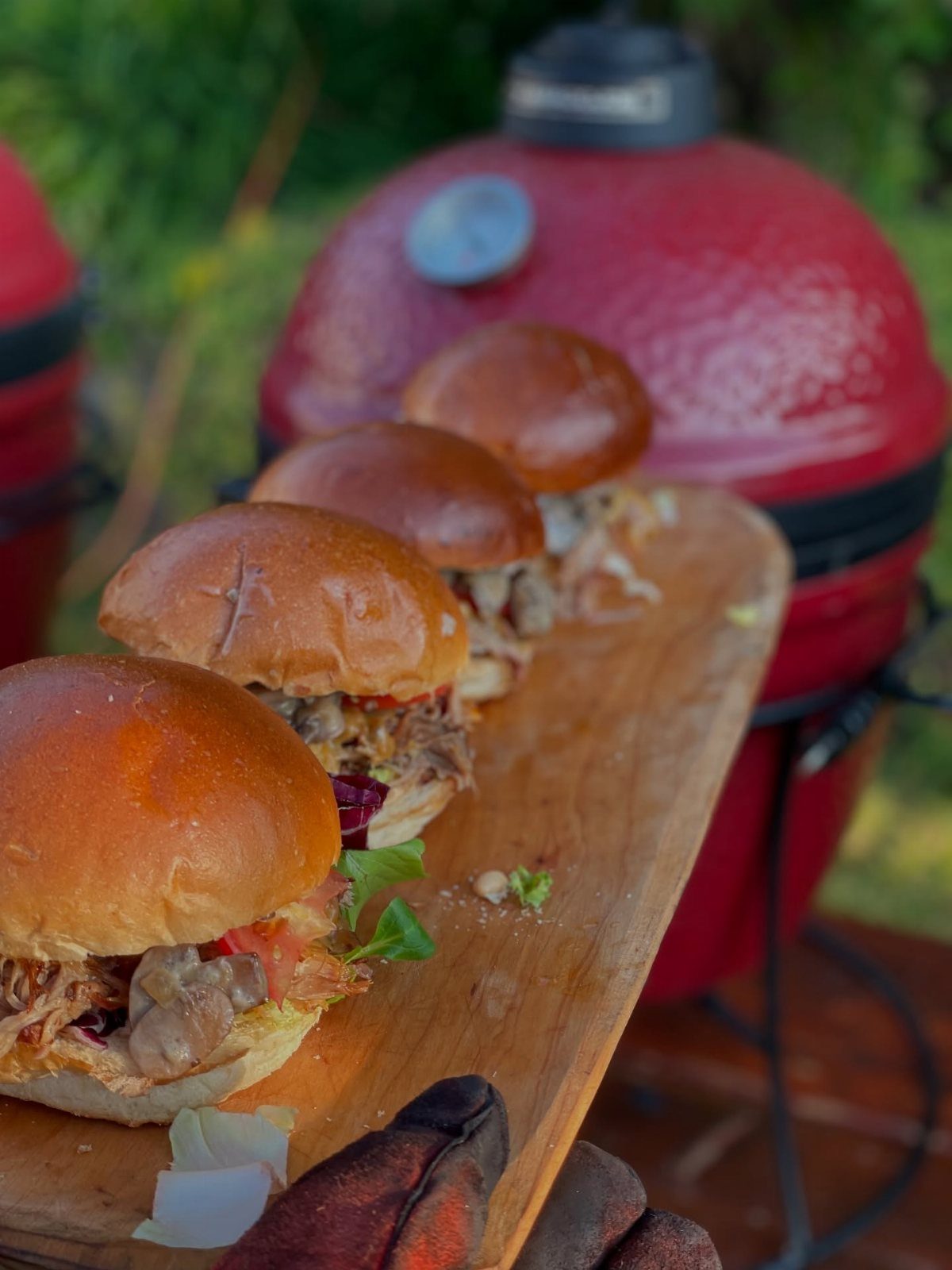 Pulled pork sliders on a wooden board with a red grill behind them.