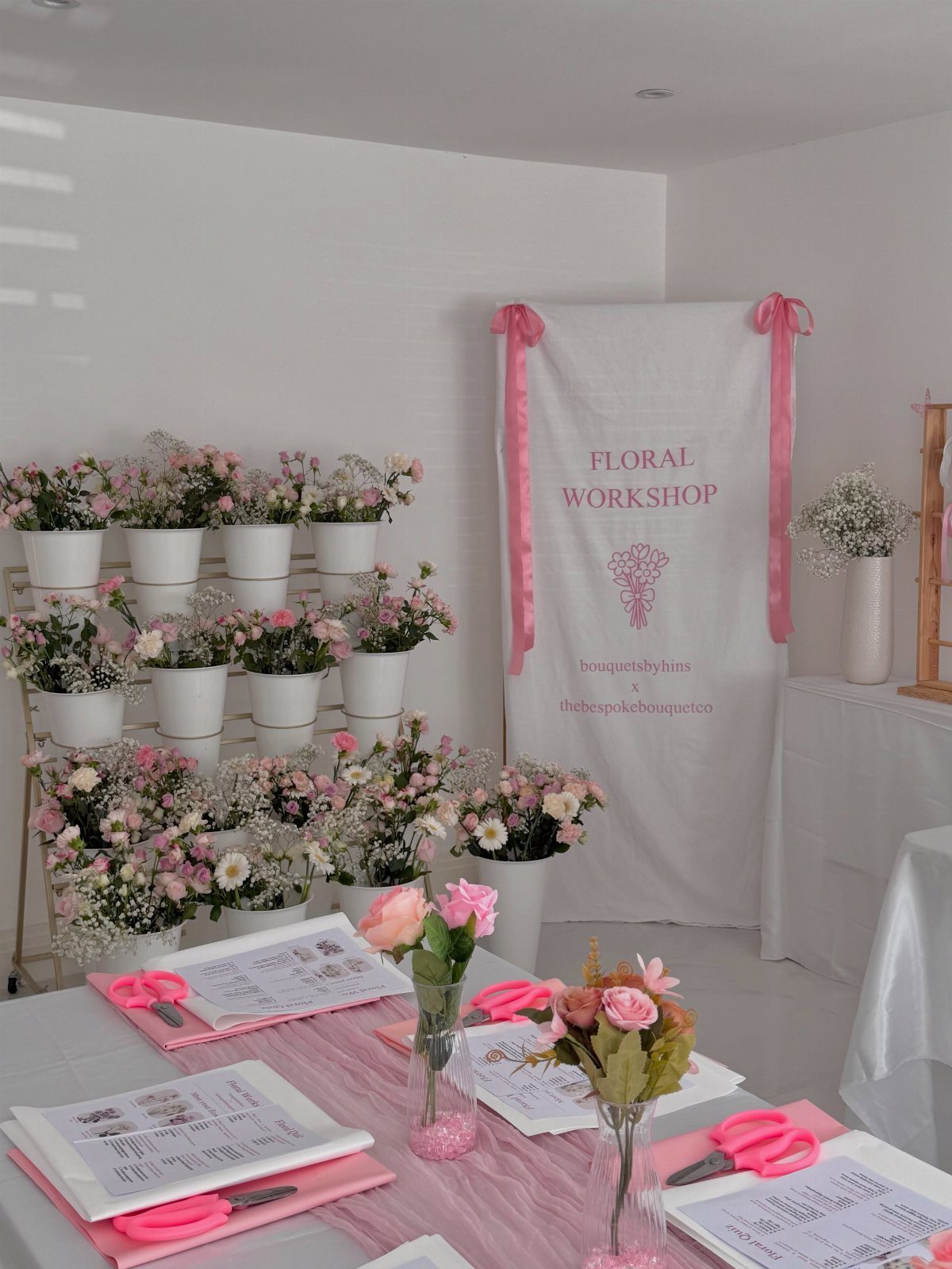 Floral workshop setup with pink flowers, white pots, and instruction sheets on a table.