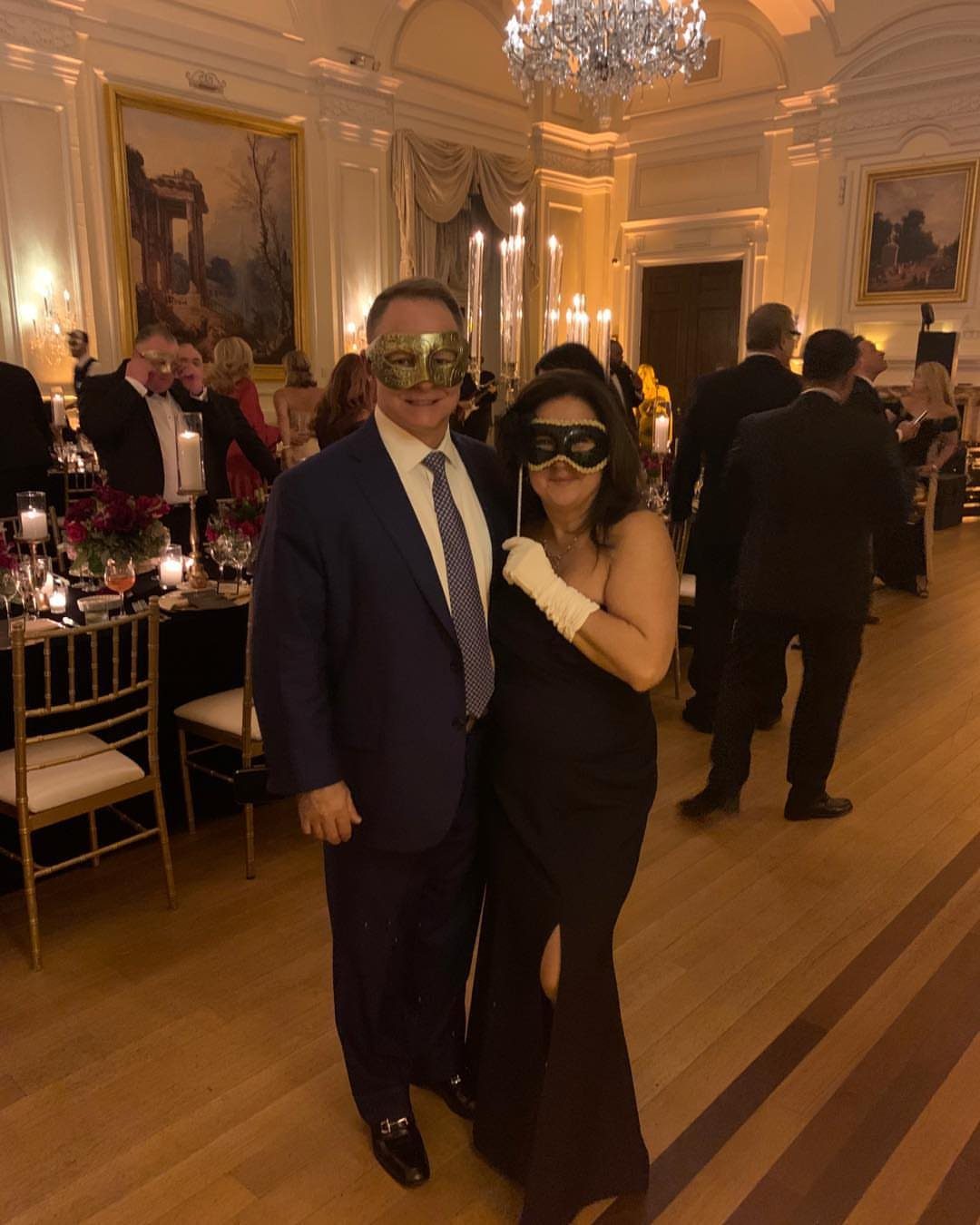 Couple wearing masquerade masks and formal attire posing in a luxury ballroom setting.