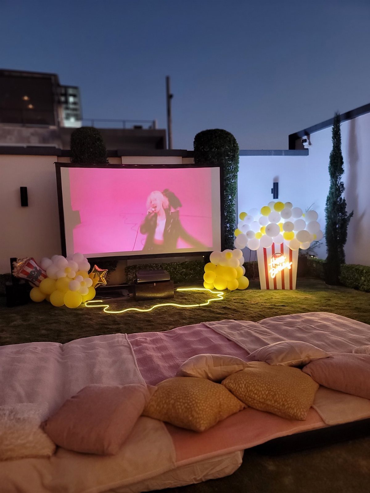 Outdoor cinema setup with a projection screen, floor mattress, and popcorn balloon decor.