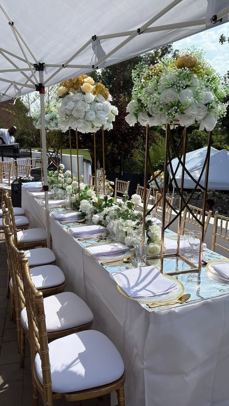 Elegant outdoor dining table with white floral centerpieces and gold chairs under a canopy