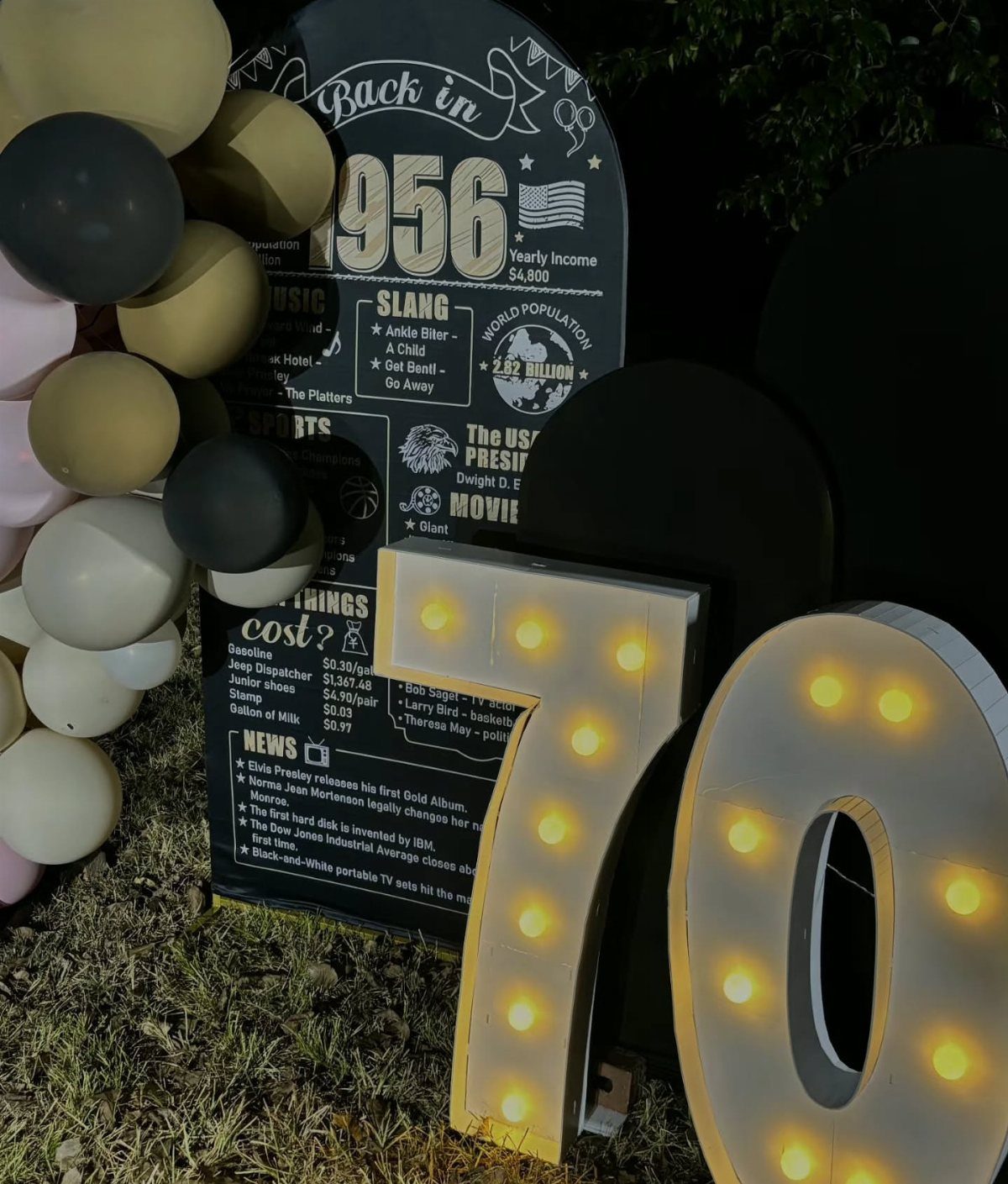 Illuminated number 70 marquee signs next to a 1956 facts board and balloons.