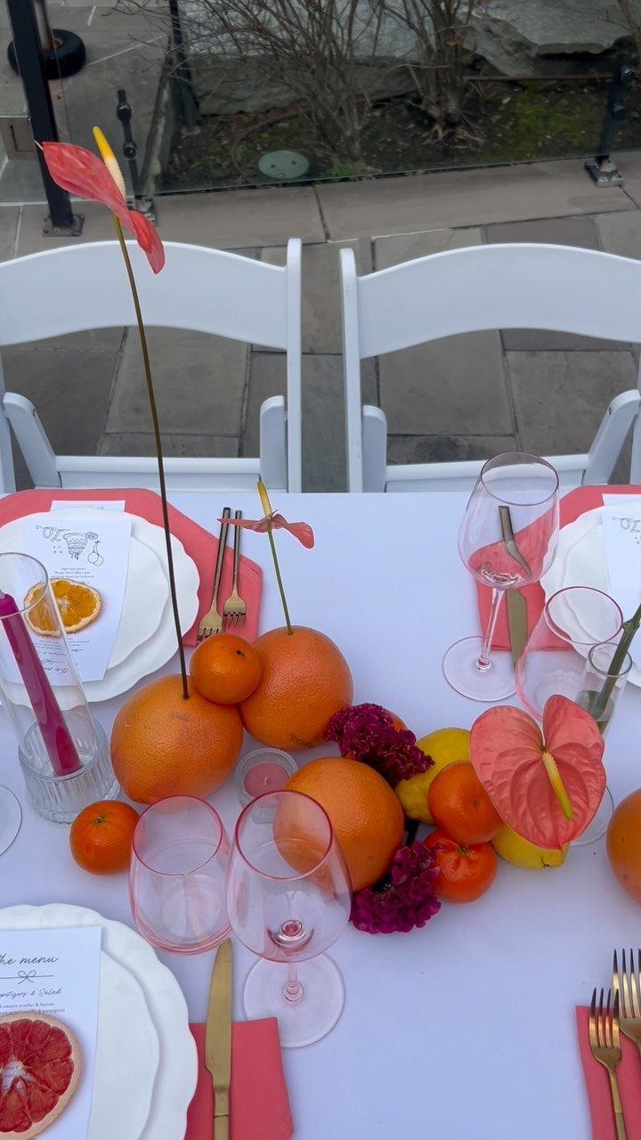 White dinner table set with pink napkins, gold cutlery, and citrus fruit centerpiece.