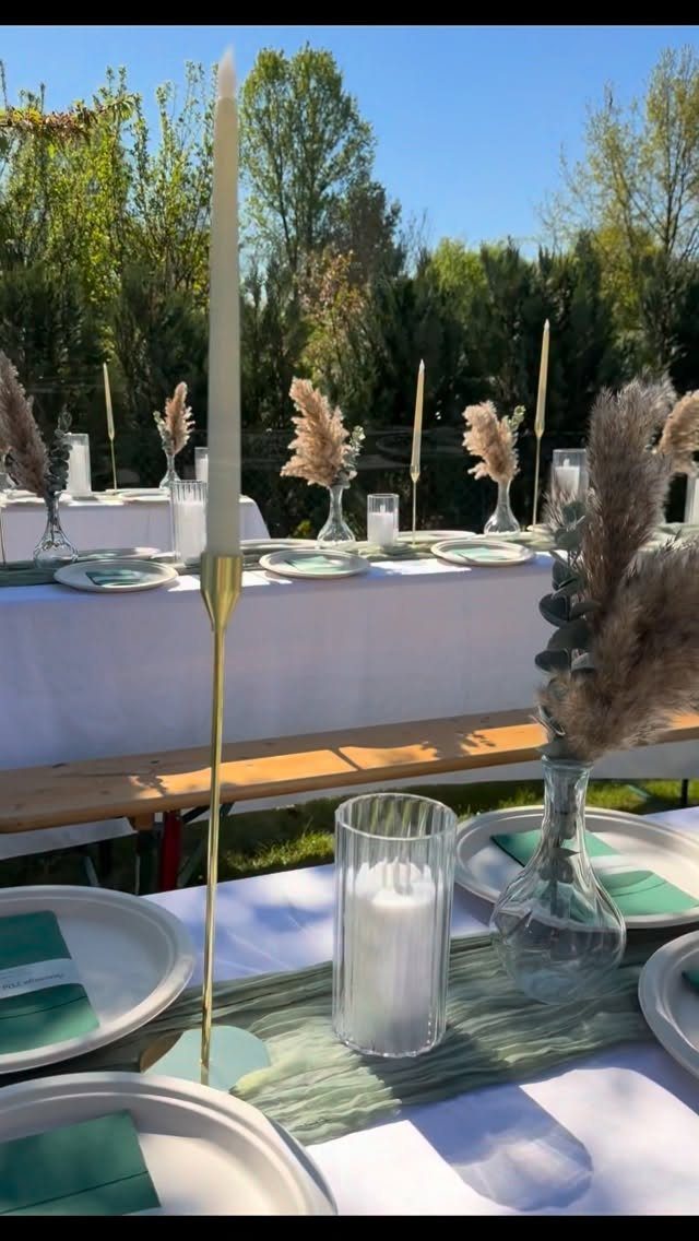Elegant outdoor table setting with white linens, green accents, and pampas grass decorations.