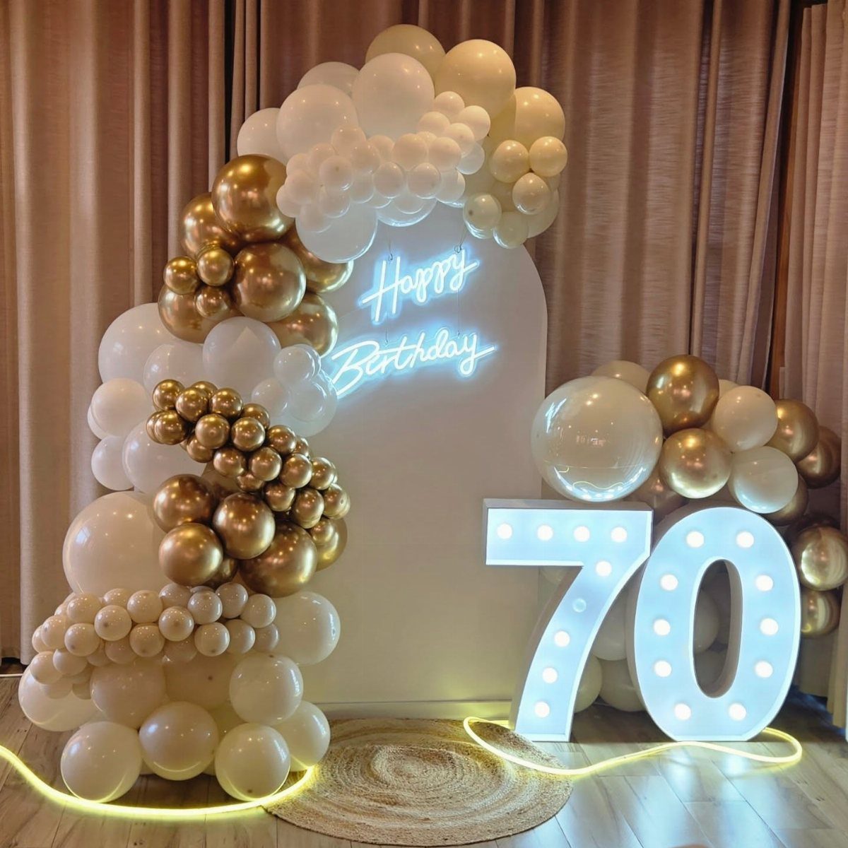 Illuminated number 70 and gold and white balloon arch for a birthday celebration.