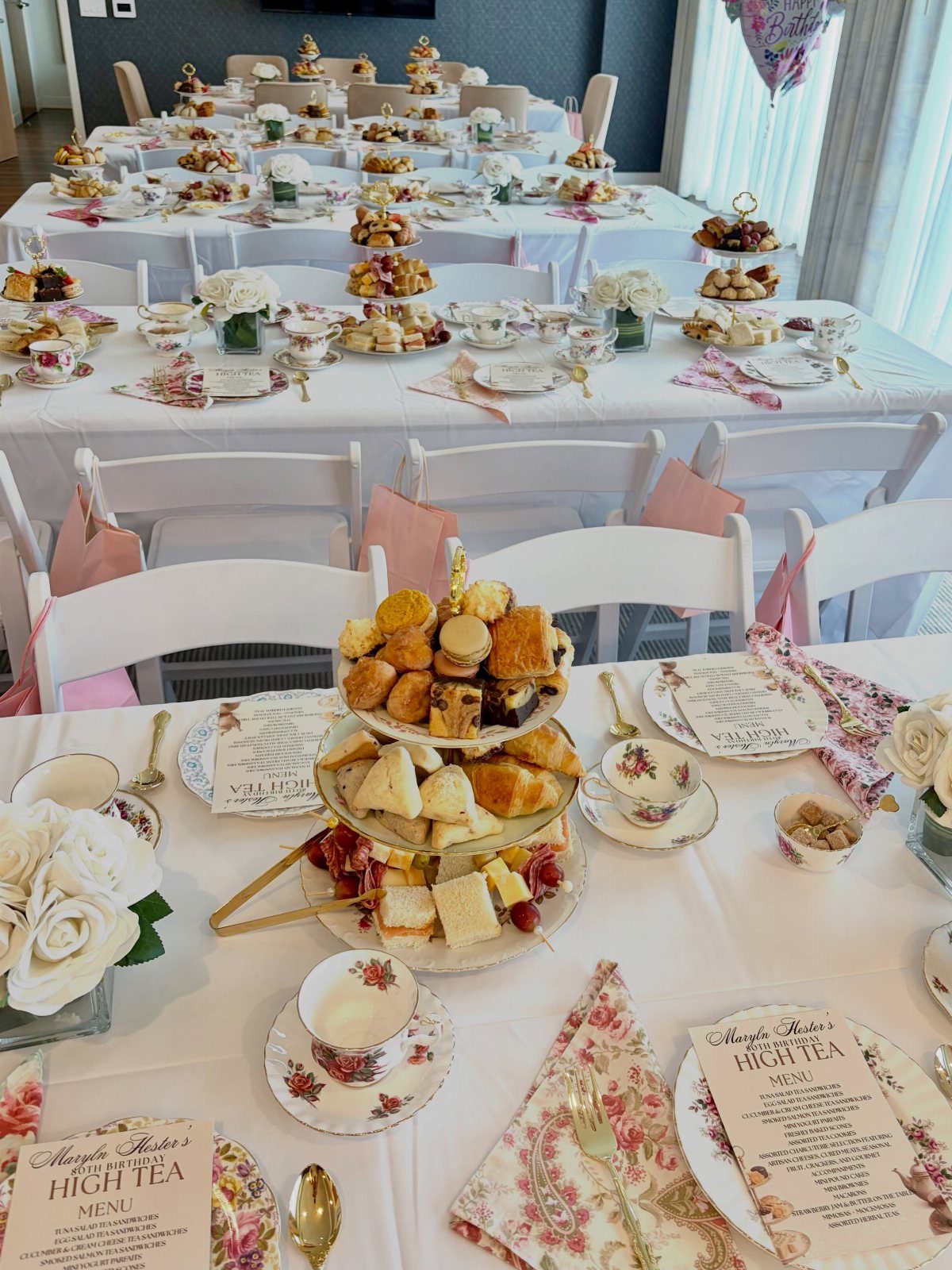 High tea table setting with tiered pastry stands, floral teacups, and white roses.