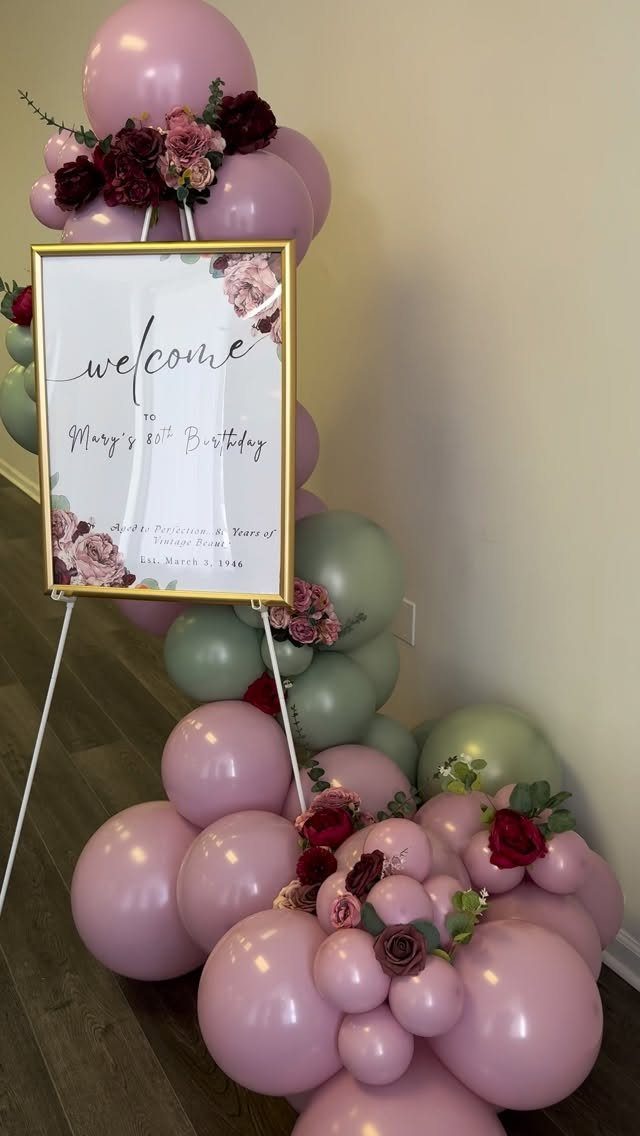 Welcome sign for 80th birthday surrounded by pink and sage green balloon arch.