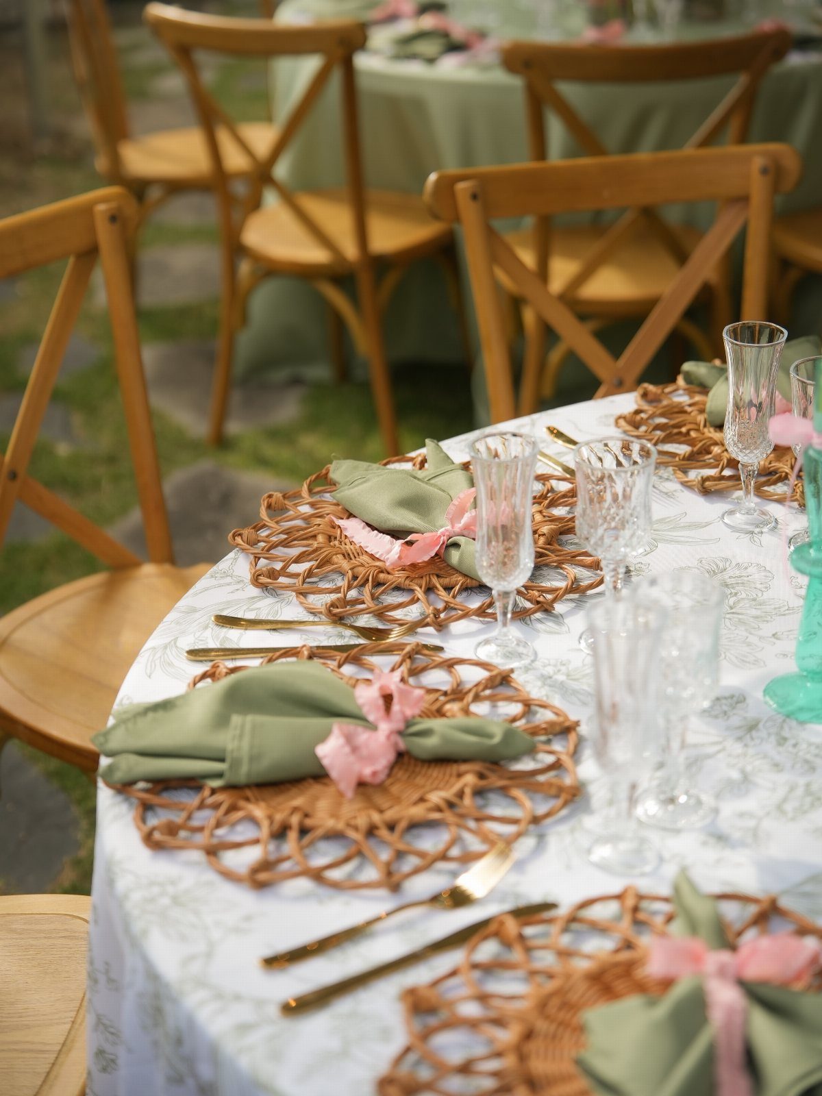 Outdoor table setting with woven placemats, green napkins, and wooden cross-back chairs.