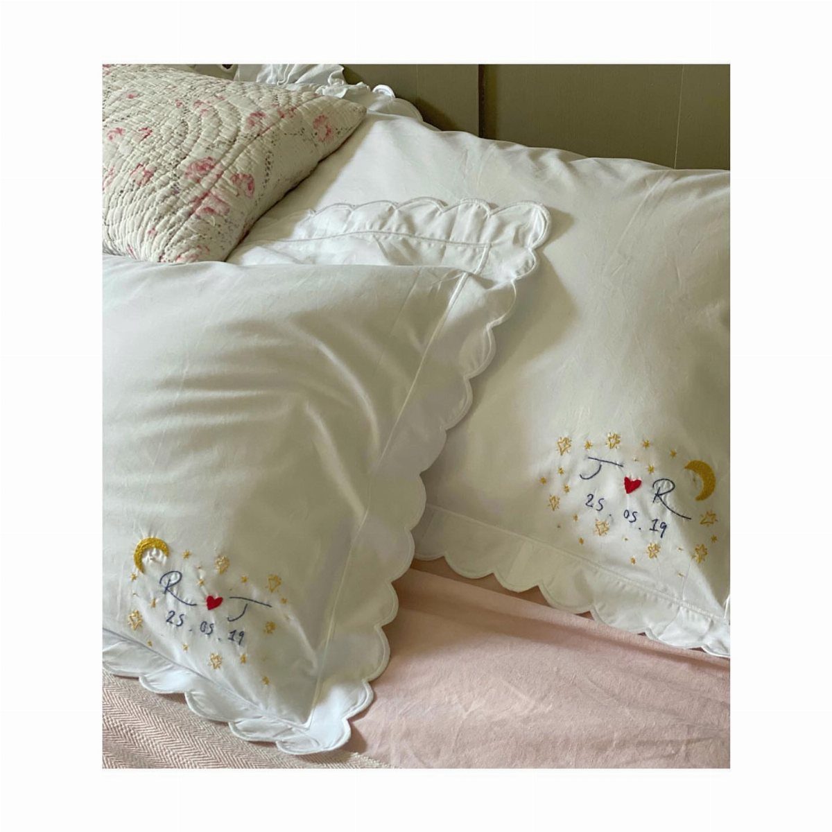 Embroidered white cotton pillowcases with initials and a date on a bed
