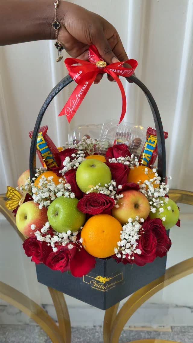 Gift basket containing red roses, white baby's breath, apples, and oranges with red ribbon.