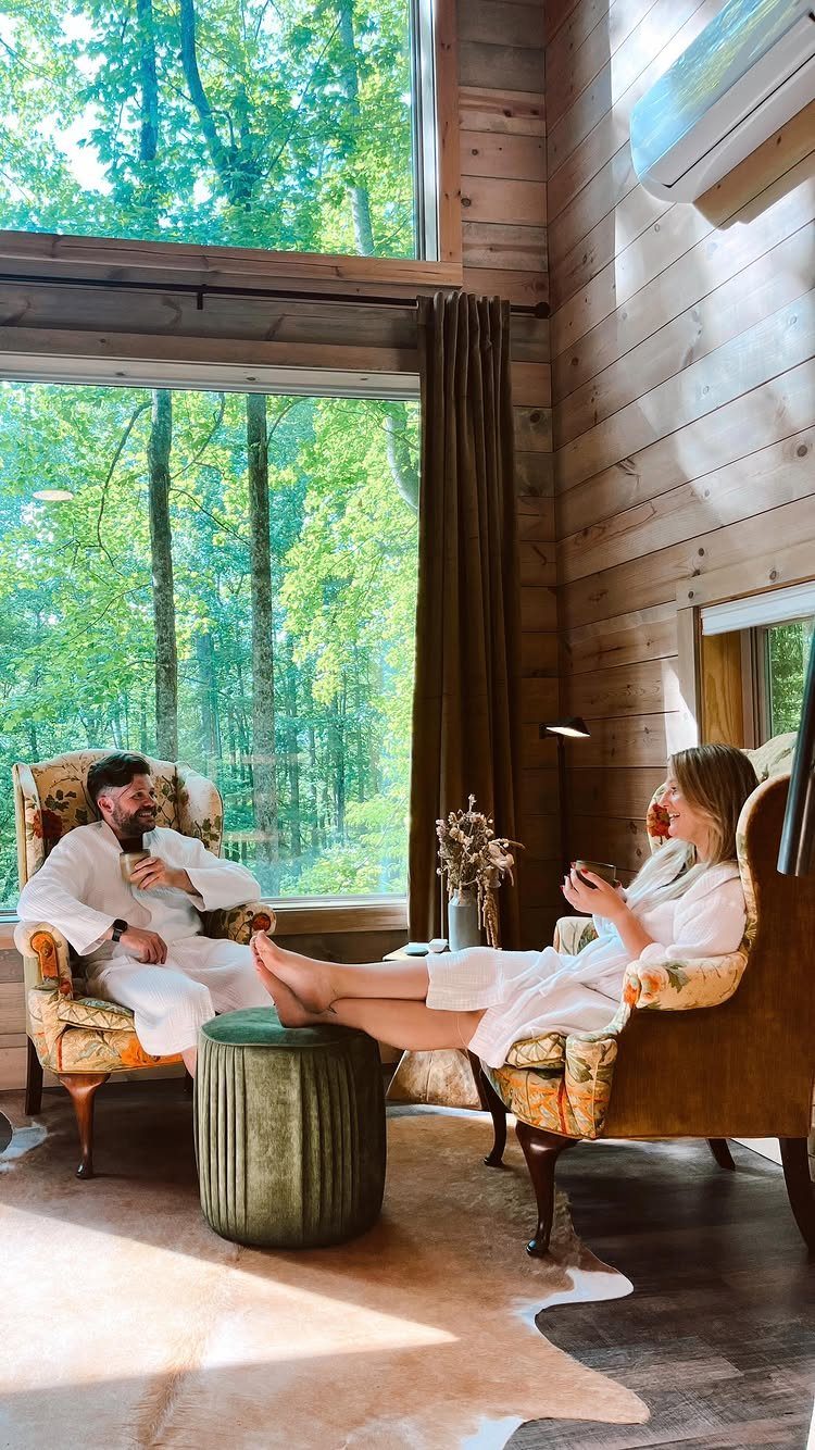 Couple in white robes relaxing in a rustic wooden cabin overlooking a forest.