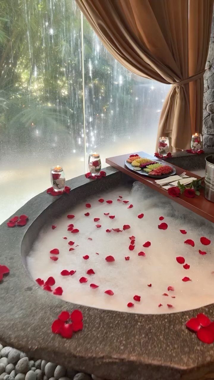 Stone bathtub with foam and red rose petals, fruit platter, and candles.