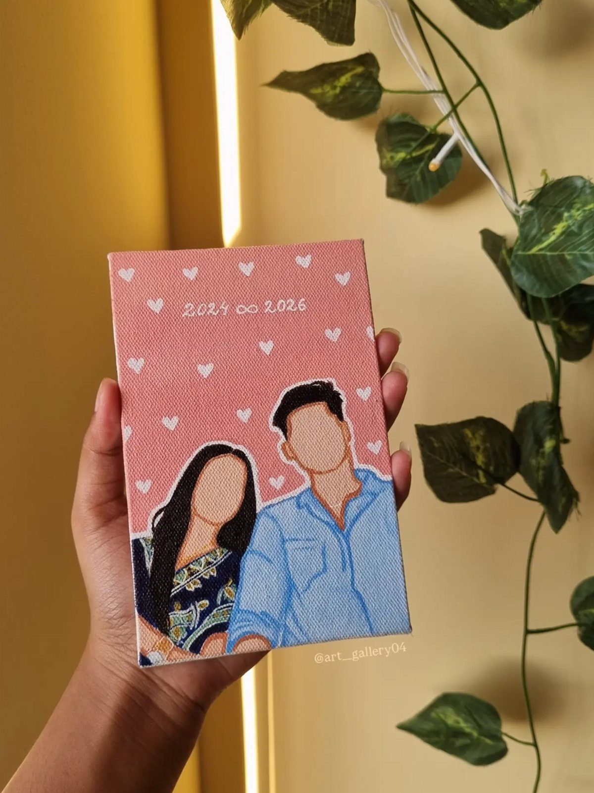 Faceless couple portrait painting on a small pink canvas held by a hand.