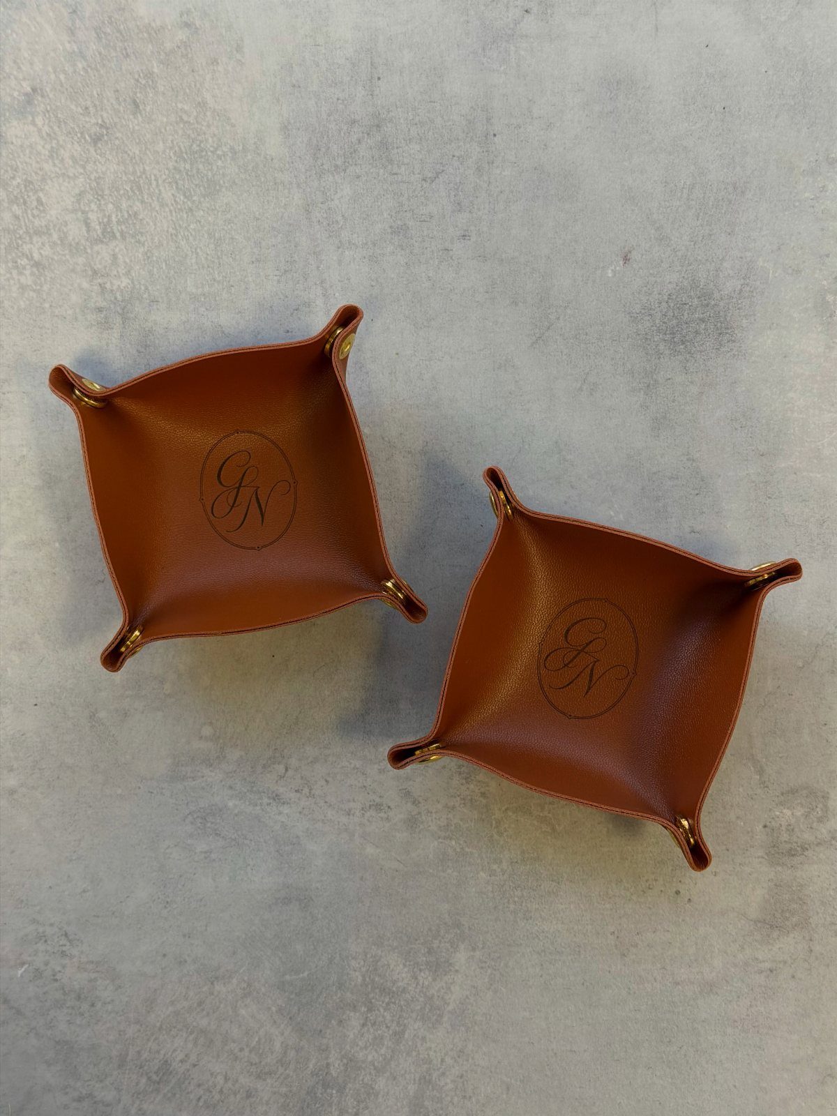 Two brown leather valet trays with embossed monograms on a grey stone background