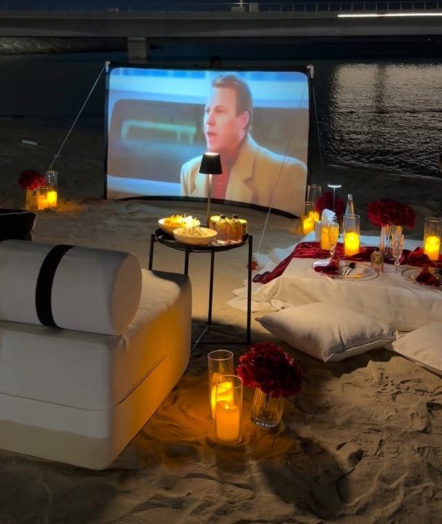 Outdoor movie screen on a beach with candles, roses, and cozy lounge seating.