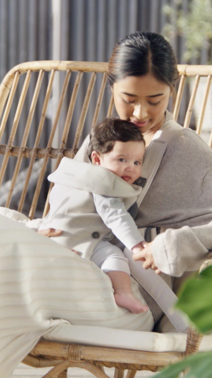 Woman sitting in a rattan chair wearing a grey baby carrier with an infant.