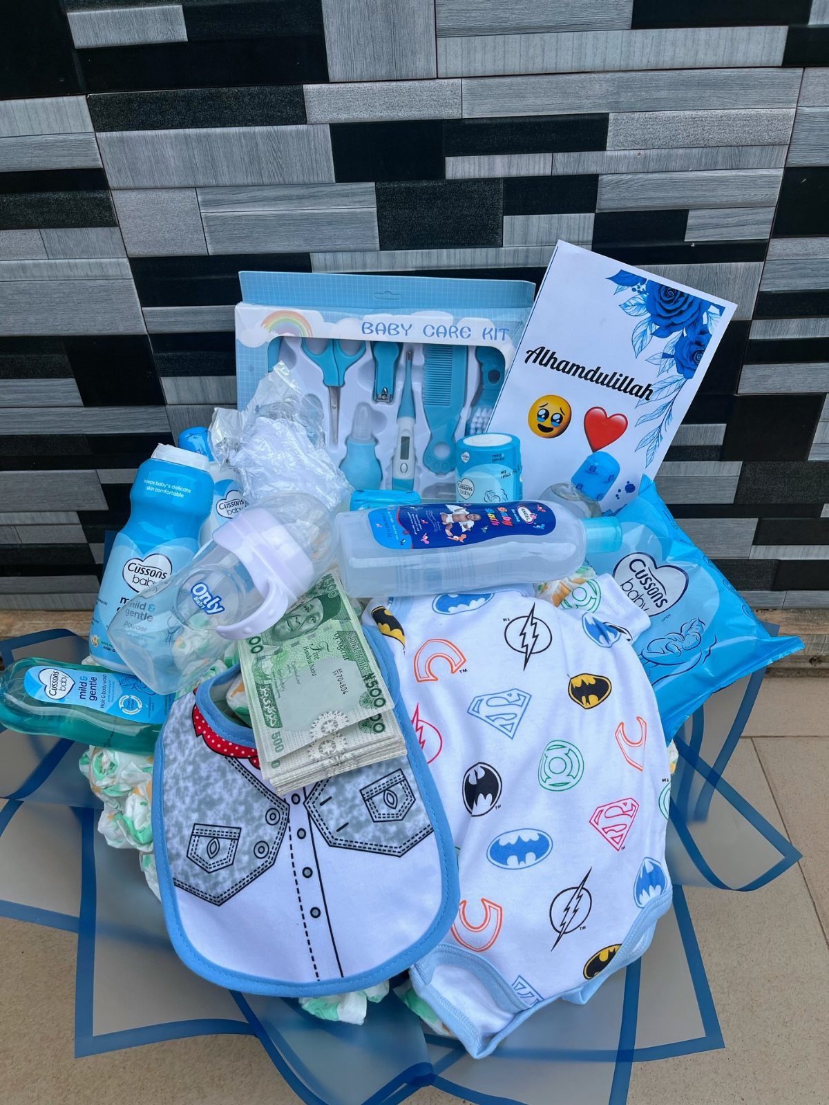 Baby gift set with superhero bodysuit, care kit, bottles, and cash on blue wrap.