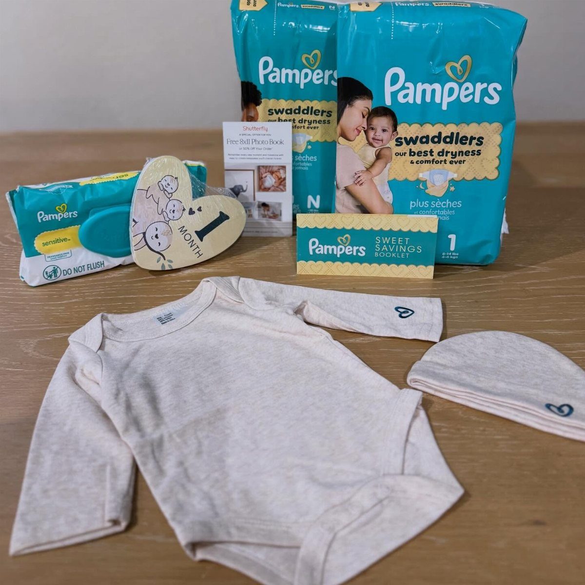 Pampers diapers and wipes with a white baby bodysuit and hat on wood.