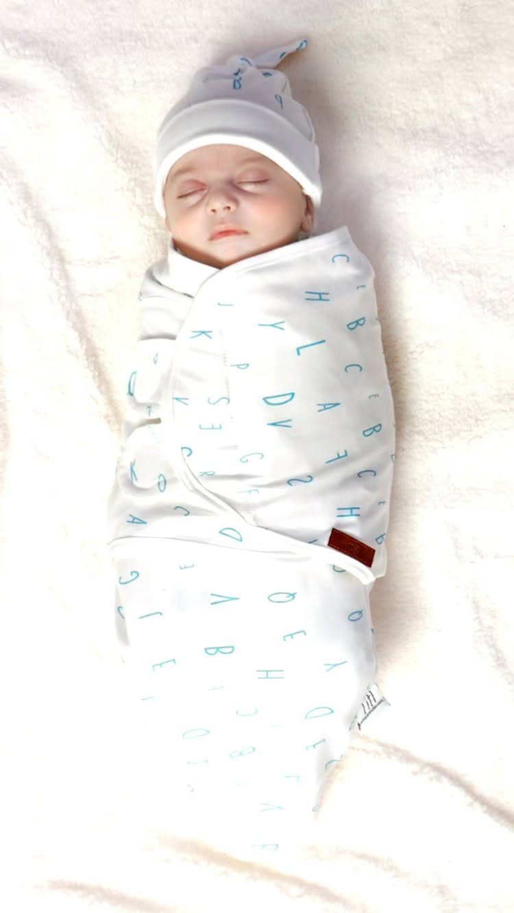Sleeping newborn baby wrapped in a white swaddle blanket with blue letters.