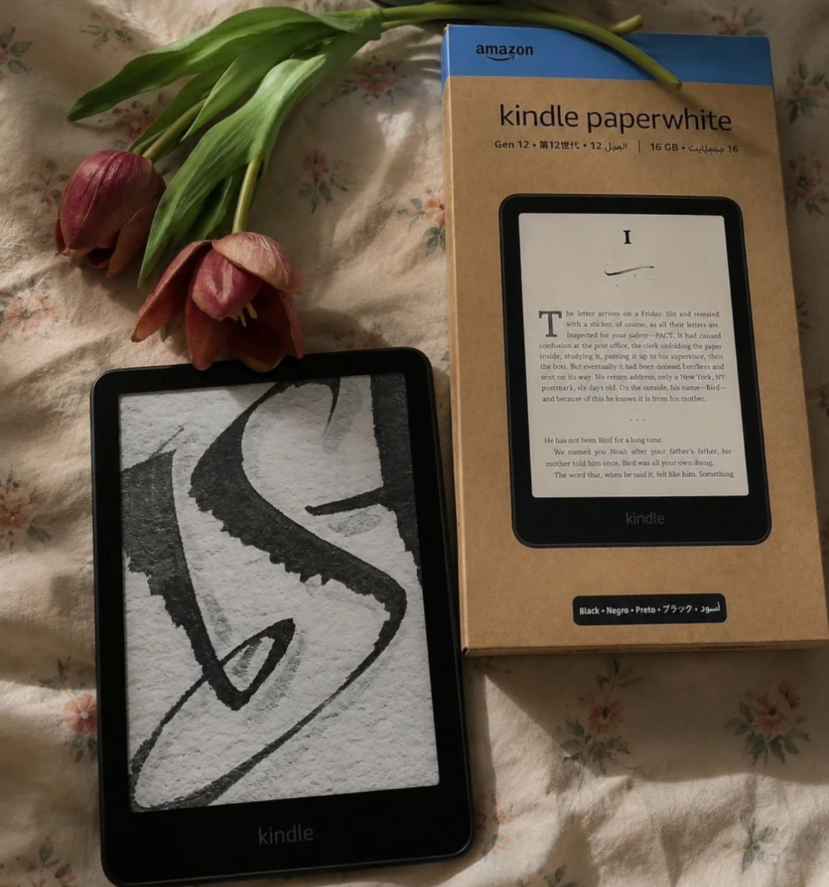 Amazon Kindle Paperwhite e-reader and its box next to red tulips on floral fabric