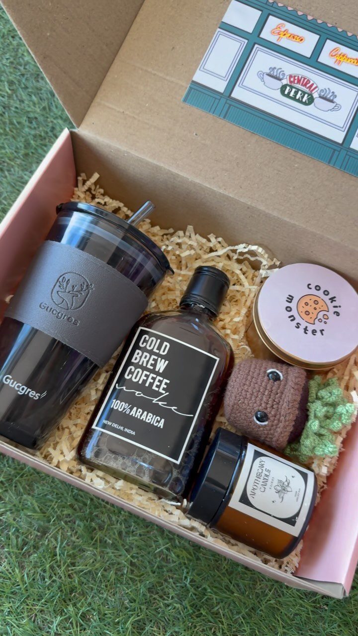 Gift box with coffee, tumbler, honey, and cookies on green grass.