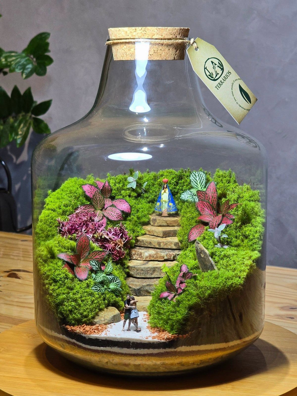 Miniature terrarium landscape in a glass jar with moss, plants, and tiny figurines.
