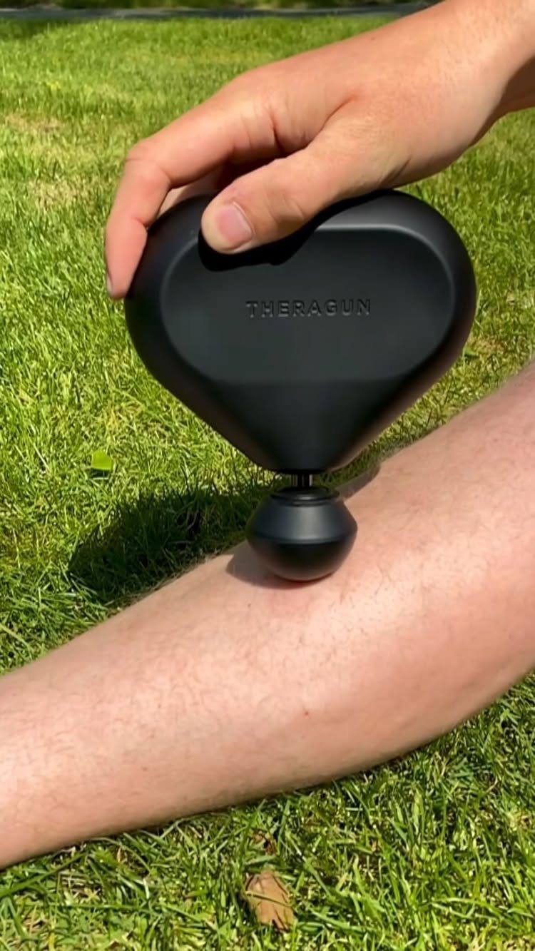 Black Theragun Mini massage gun being used on a person's leg outdoors on grass.