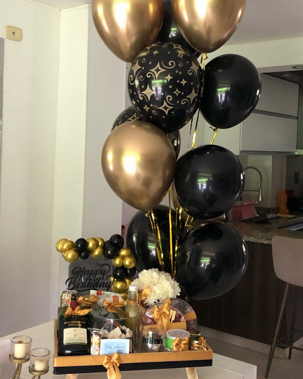 Birthday gift tray with champagne and treats next to gold and black balloons.
