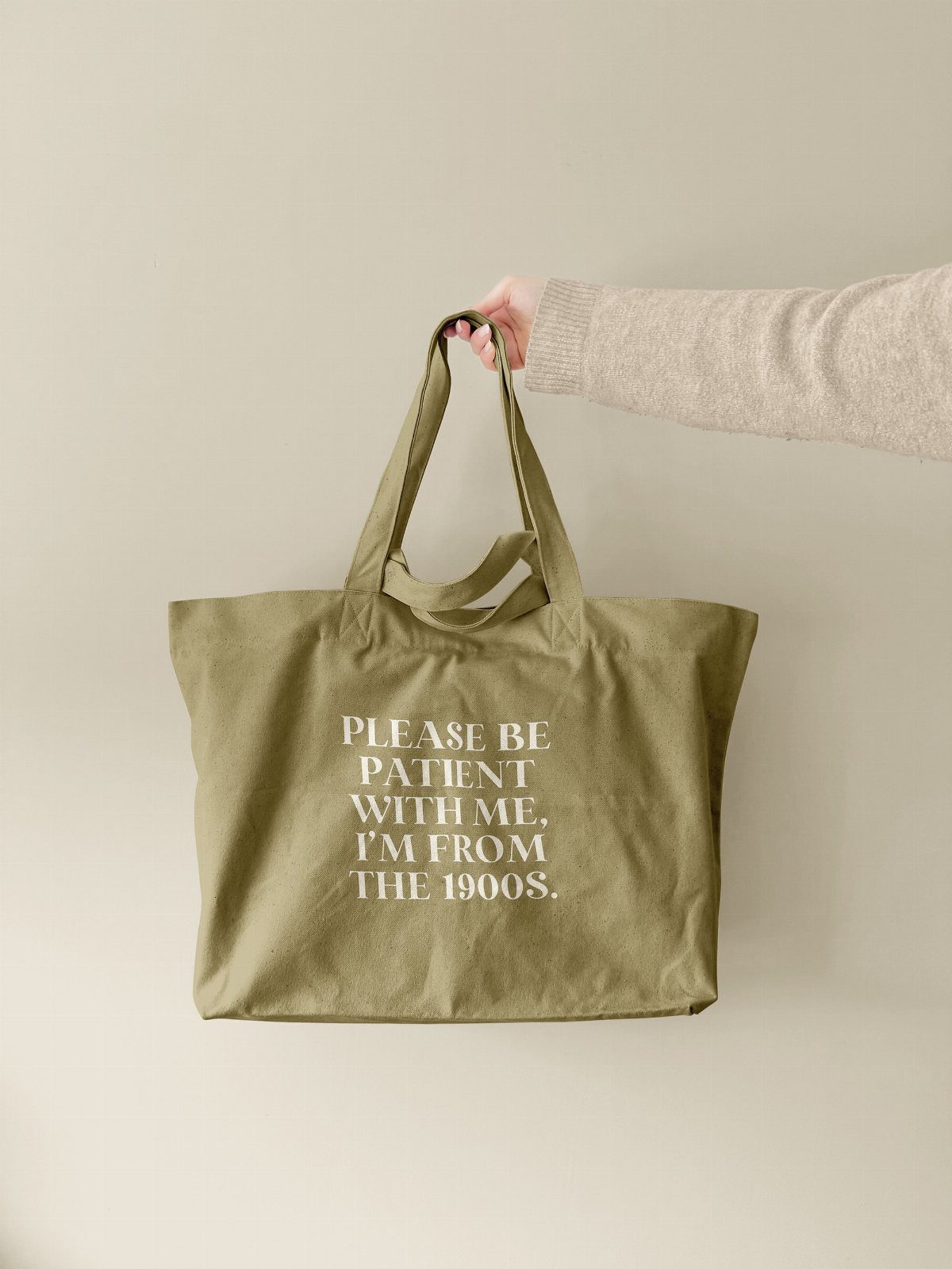 Olive green canvas tote bag with white text held against a beige wall