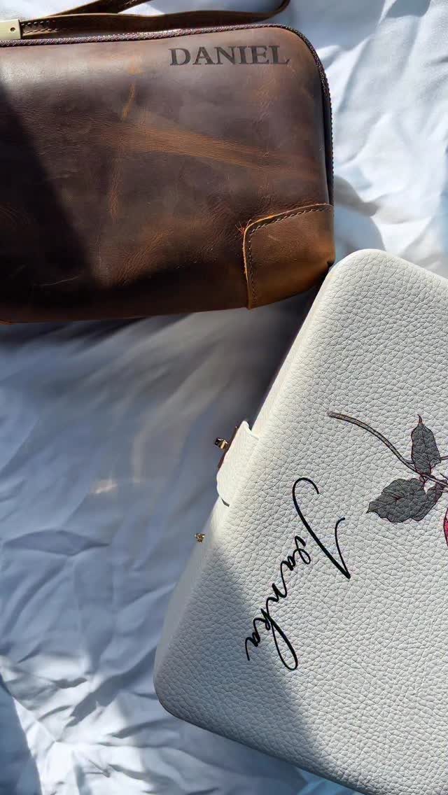 Two personalized leather toiletry bags, one brown and one white, on white fabric.