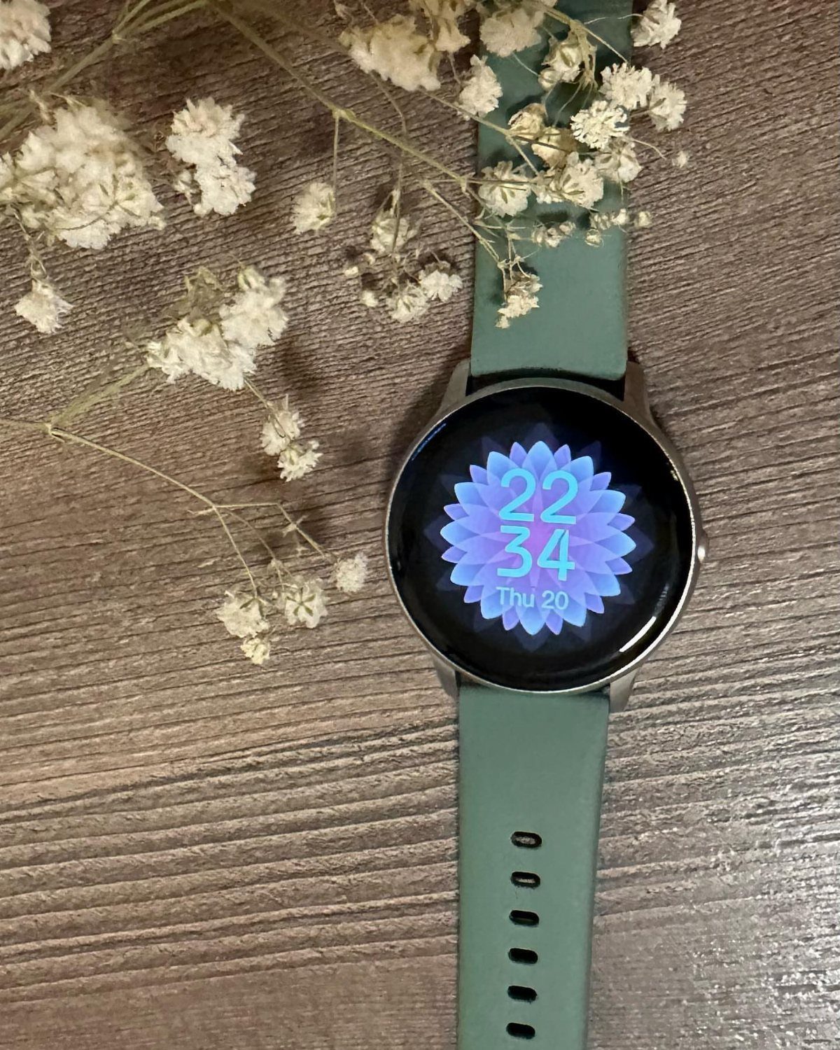 Round smartwatch with sage green strap and white dried flowers on wood.
