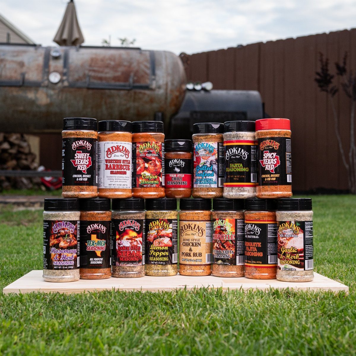 Assorted gourmet BBQ spice rub jars arranged on a wooden board outdoors.