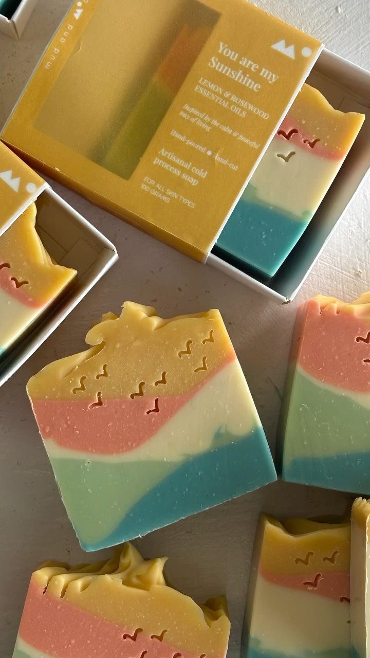 Layered yellow, pink, and teal artisan soaps with one in a yellow box.
