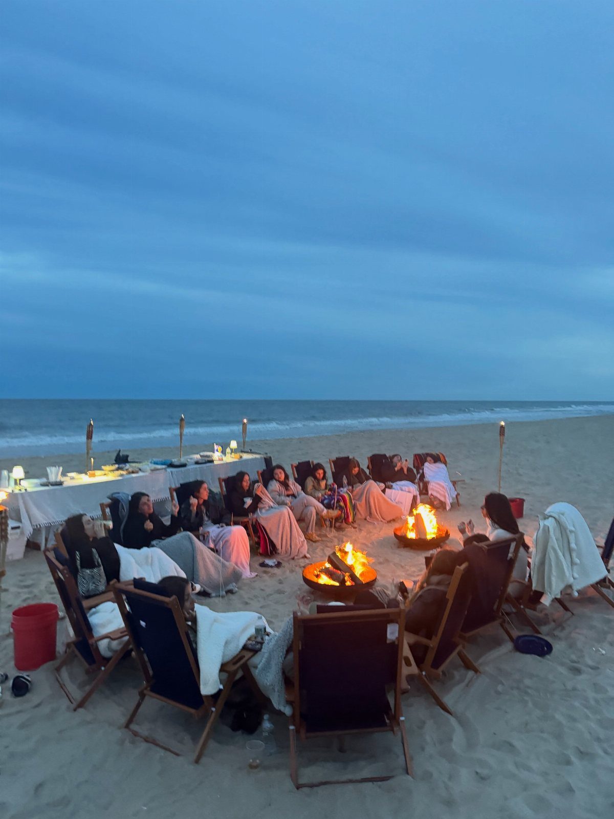 People gathered around a bonfire on a beach with a dinner table nearby.
