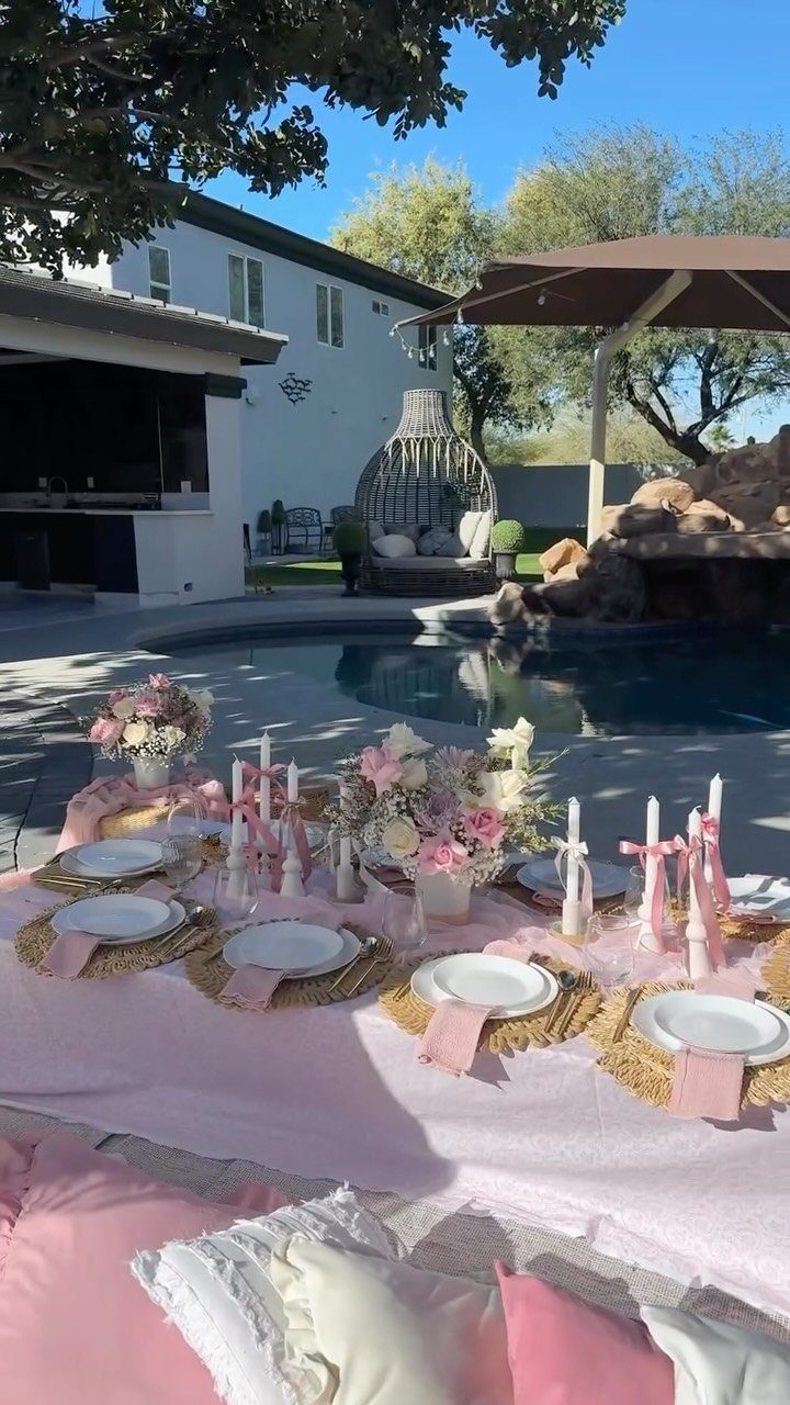 Upscale outdoor picnic setup with pink linens, white plates, and flowers by a pool.