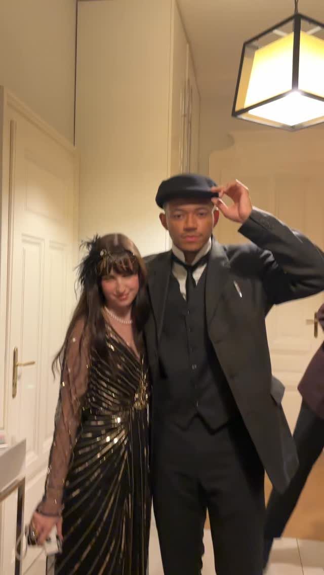 Man in a black suit and newsboy cap posing with woman in sequined dress.