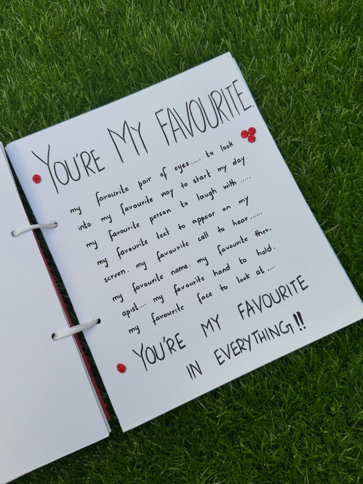 Handwritten sentimental book with red heart accents resting on a green grass background.